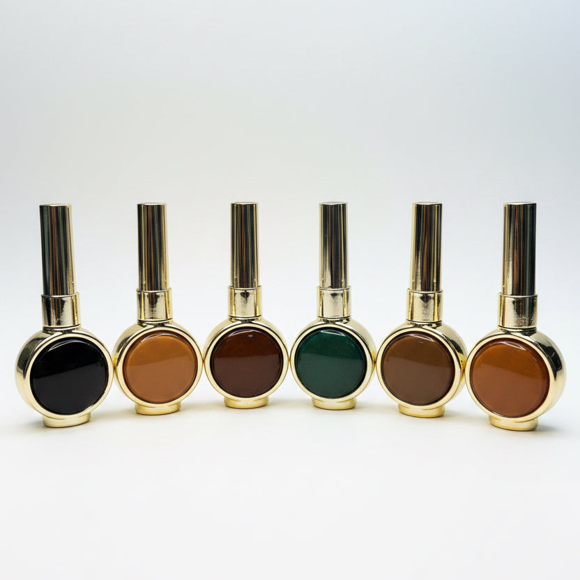 Six small cosmetic bottles with shiny gold caps and round gold-rimmed bases, arranged in a row on a wooden surface. The visible product colors in the round tops are dark brown, amber-brown, dark brown, olive green, dark brown, and amber-brown, against a plain light background.