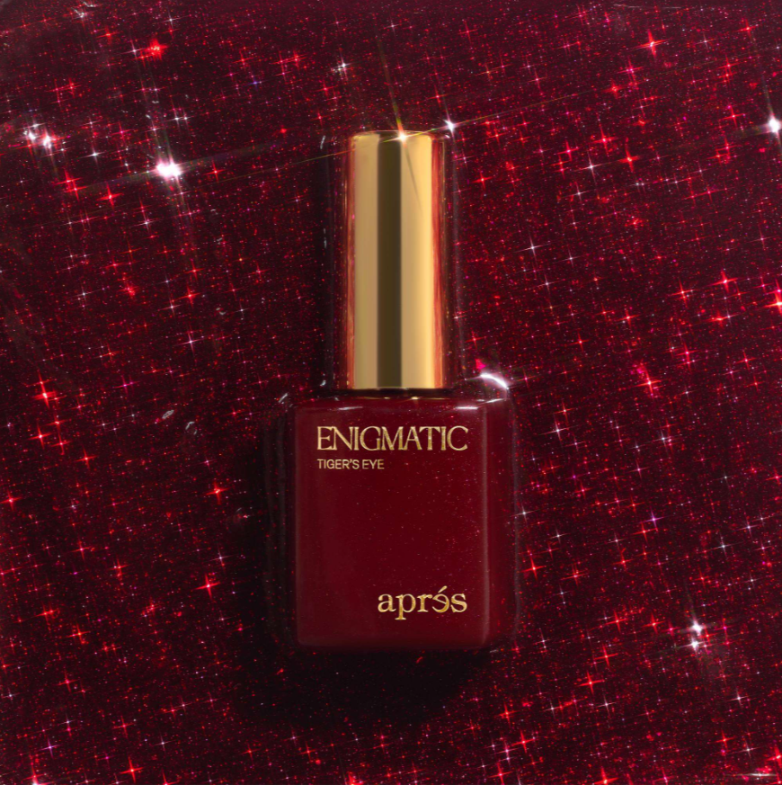 A bottle of aprés Enigmatic Tiger's Eye nail polish with a metallic gold cap, set against a sparkling red glitter background.