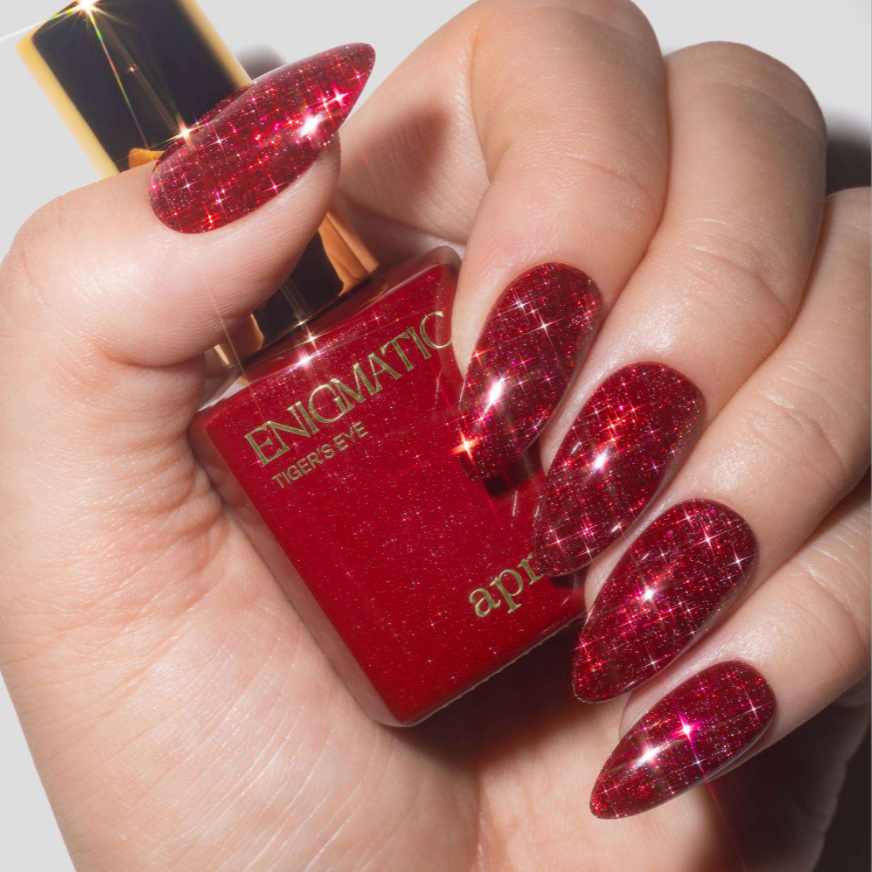 A close-up of a hand with long, glittery red manicured nails holding a bottle of Enigmatic Tigers Eye nail polish.