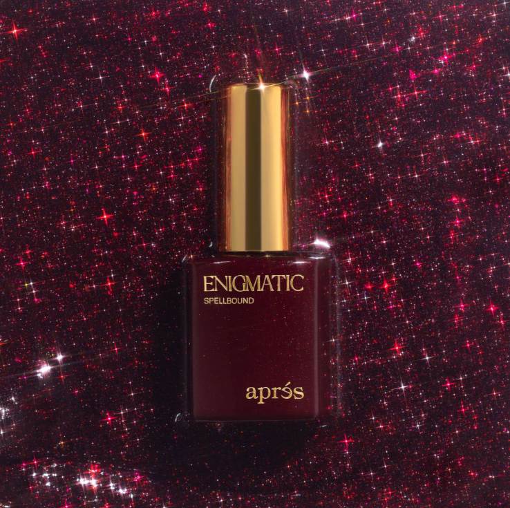 Après Enigmatic Spellbound nail polish in a dark burgundy bottle with a shiny gold cap, set against a sparkling dark red glitter background.