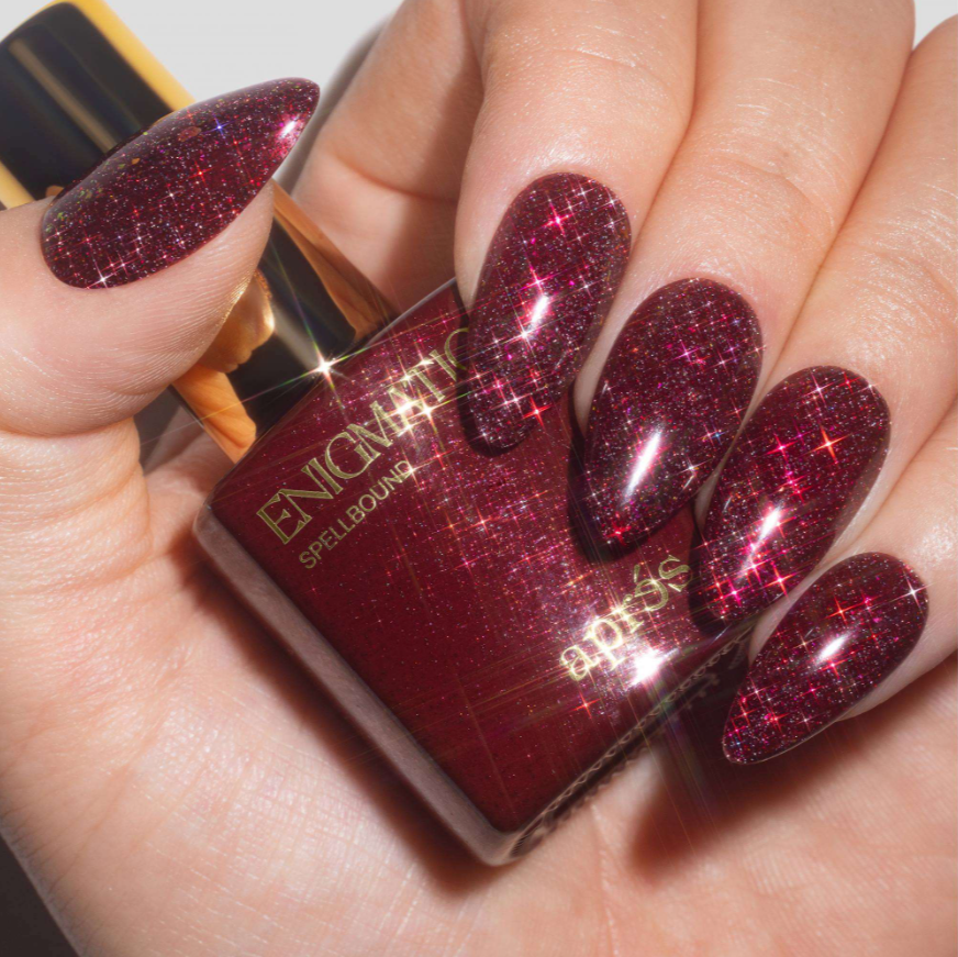 A close-up of a hand with long, stiletto-shaped nails painted in a deep burgundy glitter nail polish, holding a bottle of the same glitter polish. The bottle is labeled 'ENCHANTED SPELLBOUND' and 'apothecary'.