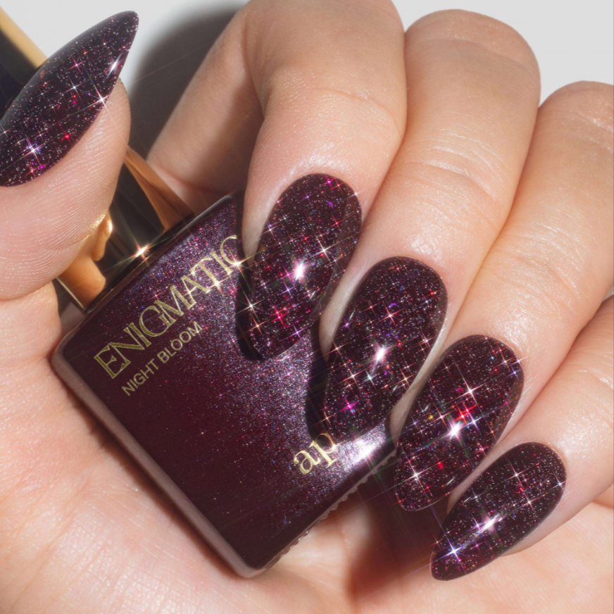 A close-up of a hand with long almond-shaped nails painted in a deep burgundy glitter nail polish with pink and purple sparkles, holding a bottle labeled ENOMATIC Night Bloom.