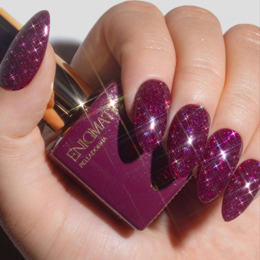 A close-up of a hand with long, glittery magenta-purple almond-shaped nails holding a bottle of nail polish. The bottle label reads ENCIWALKER RELIGION.