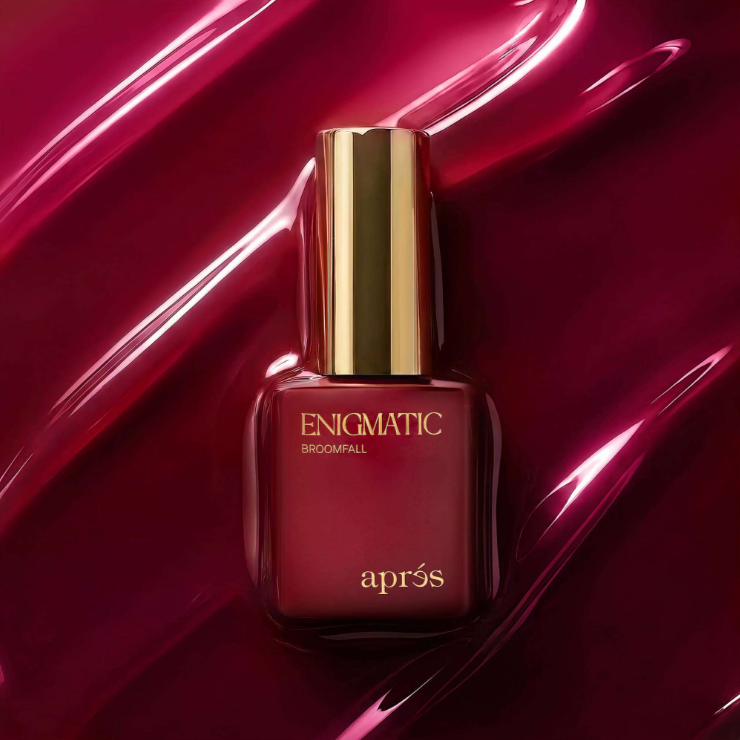 Apres Enigmatic Broomfall nail polish in a dark red bottle with a shiny gold cap, set against a glossy, dark red liquid background.