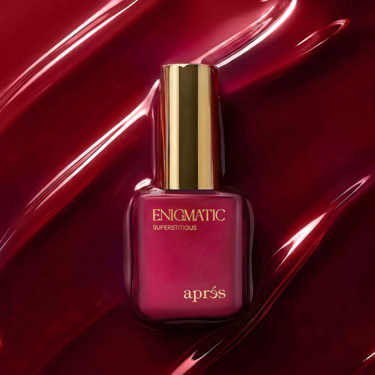 Apres Enigmatic Superstitious nail polish in a deep burgundy shade, presented in a square glass bottle with a shiny gold cap, set against a glossy, abstract red background.