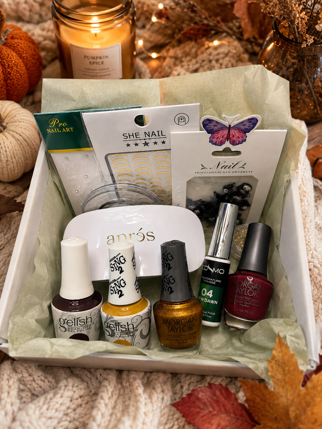 A fall-themed nail care gift box, containing three nail polish bottles, a white nail file, a sheet of gold nail art, and a butterfly-shaped press-on nail pack, arranged in a white box lined with light green tissue paper on a cozy knit blanket.