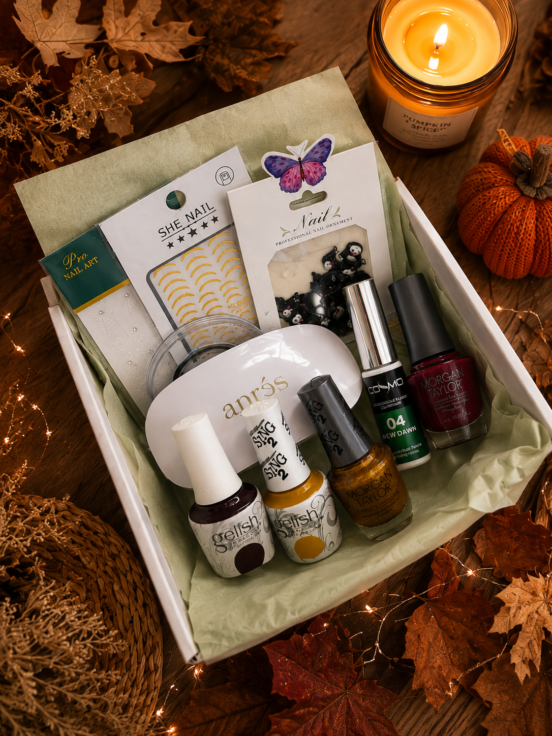 A curated fall-themed gift box containing nail care and beauty items, including an anvil-shaped nail file, multiple nail polish bottles, a Shell Nails set, and a butterfly nail art sticker, arranged in a white box with green tissue paper on a rustic wooden table surrounded by autumn leaves, a candle, and a small pumpkin.