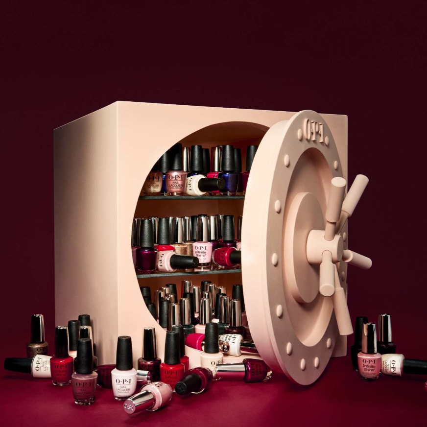 A beige novelty safe styled as a nail polish collection display, with shelves inside filled with colorful nail polishes and additional bottles arranged around it on a dark burgundy background.