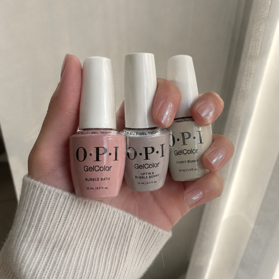 A hand holding three OPI GelColor bubble bath scented nail polishes in soft pink, white, and light gray shades, set against a soft beige curtain background.