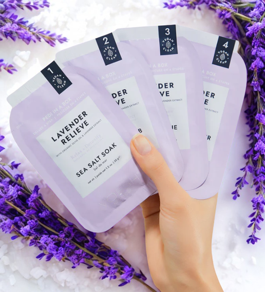 Hand holding four lavender-themed packaging pouches with lavender flowers in the background