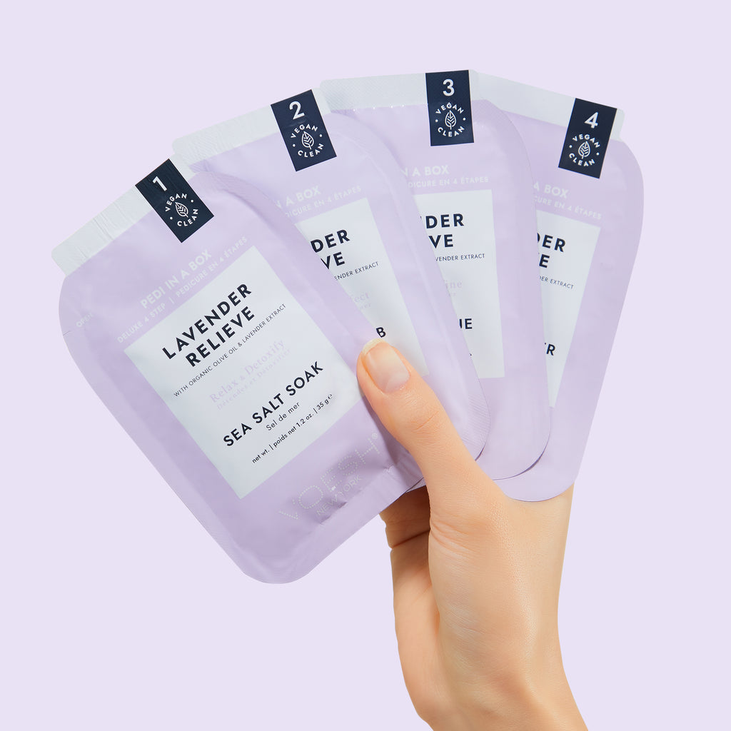 Hand holding four lavender sea salt soak packages against a light purple background