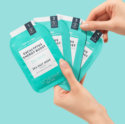 Hand holding four Voesh Eucalyptus Energy Boost sea salt soaks against a light blue background