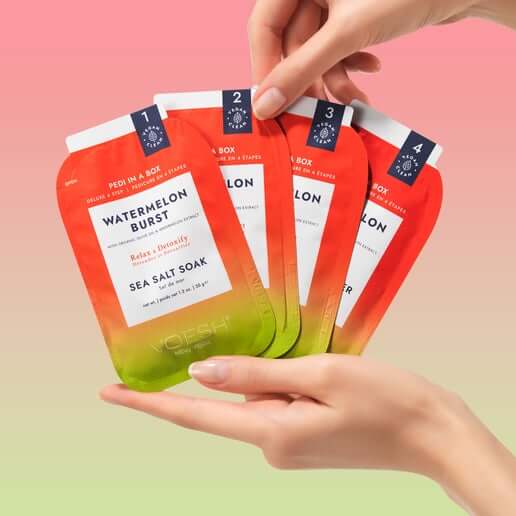 Hand holding four packets of 'Watermelon Burst' sea salt soaks with a gradient design on a pink and green background.